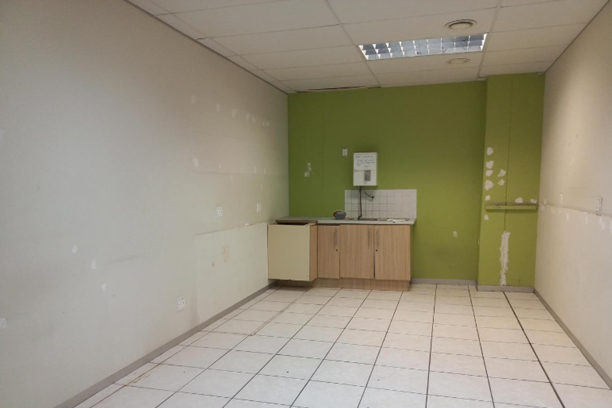 An empty office kitchenette area featuring basic cabinetry, a sink, and tiled flooring, requiring cosmetic updates and wall repairs.