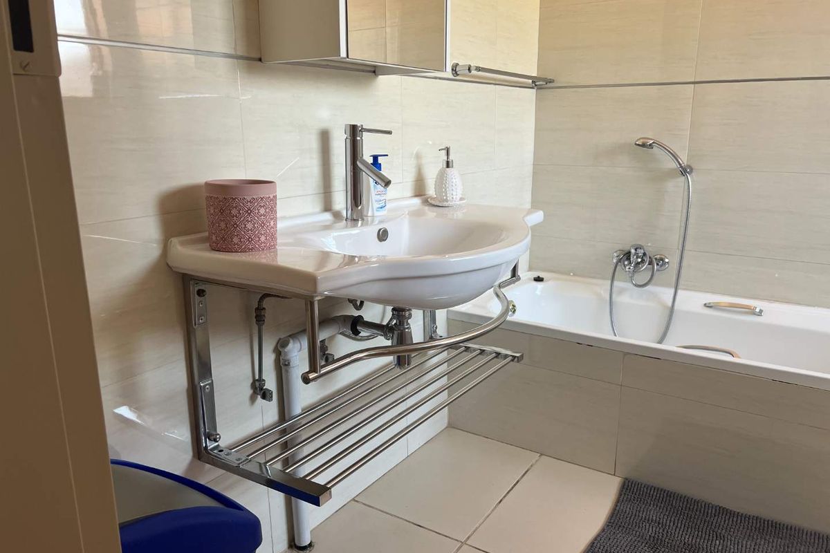A functional bathroom featuring a wall-mounted basin with integrated towel rack, a bathtub with handheld shower attachment, and neutral wall tiling.