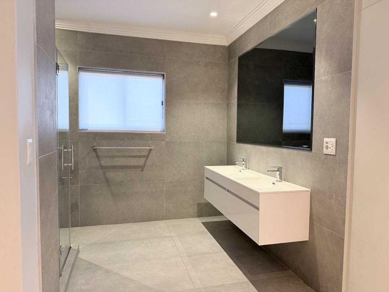 Modern bathroom featuring a sleek wall-mounted double vanity, large mirror, glass shower enclosure, and contemporary grey floor-to-ceiling tiling.