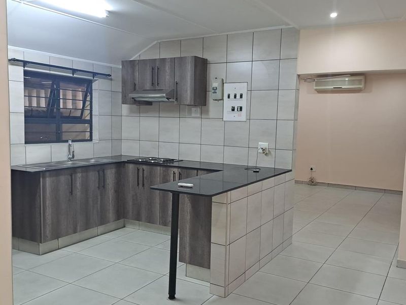 A clean, modern kitchen featuring dark wood-look cabinetry, black countertops, tiled splashbacks, and an integrated breakfast bar, flowing into an open-plan living area.