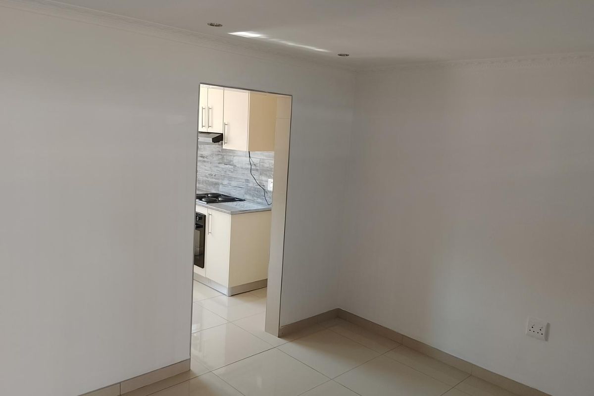 A bright, empty living area with neutral tiled flooring, featuring a direct view into a modern kitchen with built-in cabinetry and appliances.