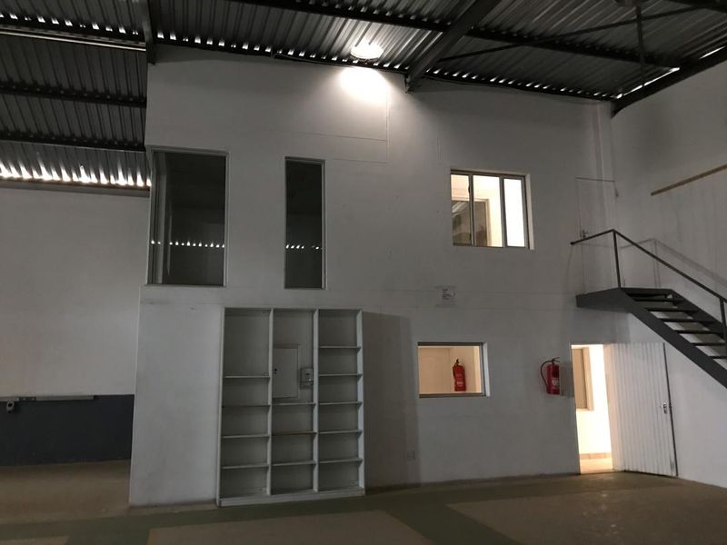 Industrial warehouse interior featuring a steel staircase, built-in shelving, and mezzanine-level office windows under a high corrugated metal roof.