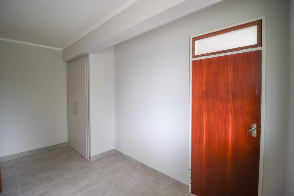 A clean, empty bedroom featuring neutral tiled flooring, a built-in wardrobe, and a classic wooden door with a transom window.