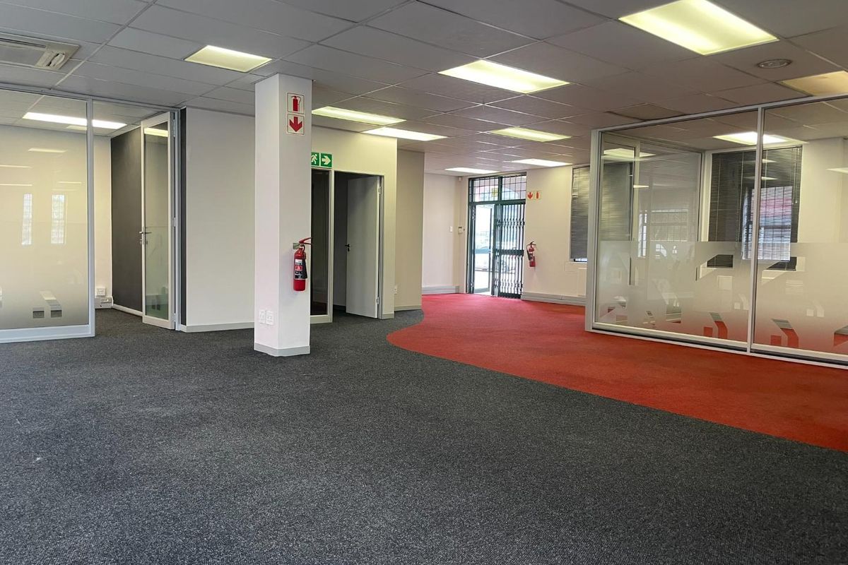 Spacious open-plan office area featuring glass partitioned rooms, suspended ceilings with recessed lighting, and dual-tone carpet flooring.