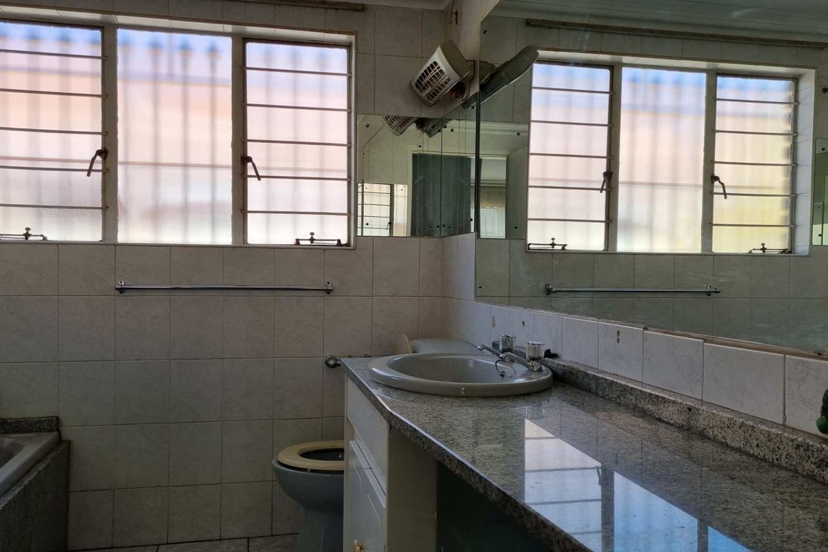 A dated bathroom featuring a granite vanity top, integrated basin, bathtub, and large wall-mounted mirror with tiled walls and security bars on windows.