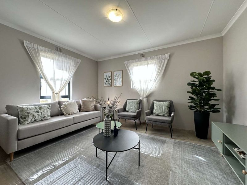 A well-lit living room featuring neutral wall tones, tiled flooring, and dual windows providing natural light.