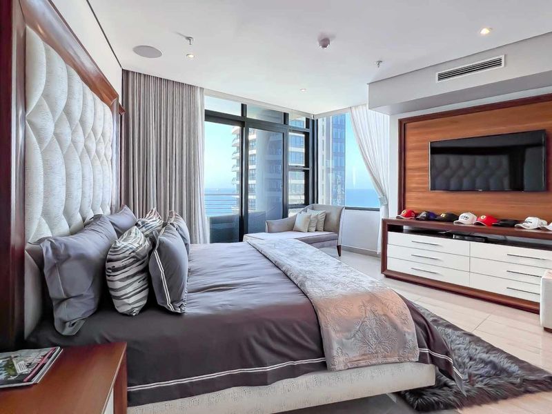 Spacious modern bedroom featuring floor-to-ceiling windows with ocean views, integrated ceiling speakers, ducted air conditioning, and custom wood-paneled media wall.