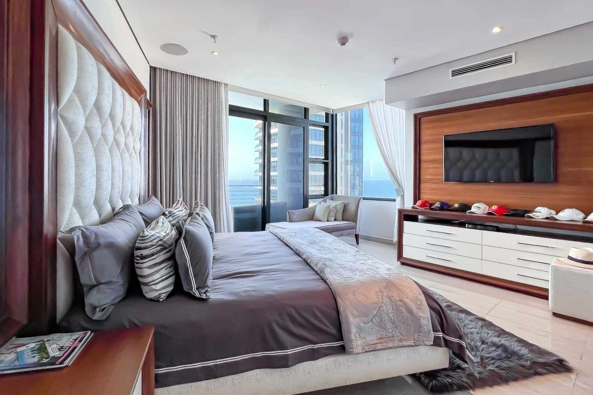 Spacious modern bedroom featuring floor-to-ceiling windows with ocean views, integrated ceiling speakers, ducted air conditioning, and custom wood-paneled media wall.