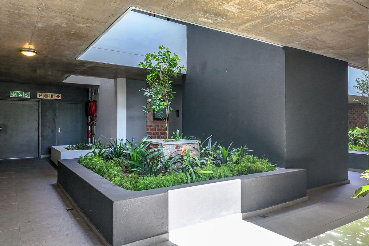 A modern, semi-enclosed communal courtyard featuring exposed concrete ceilings, tiled flooring, and a central landscaped planter box with integrated lighting.