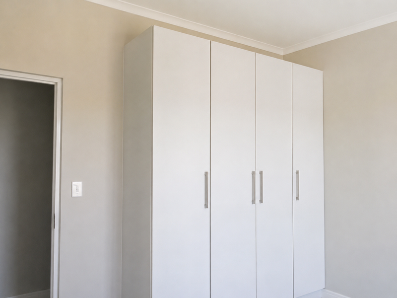 A clean, neutral bedroom featuring a modern white built-in wardrobe with sleek metal handles and simple coving.