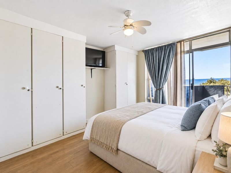 A bright bedroom featuring built-in cupboards, a ceiling fan, and a sliding glass door that opens to a balcony with ocean views.
