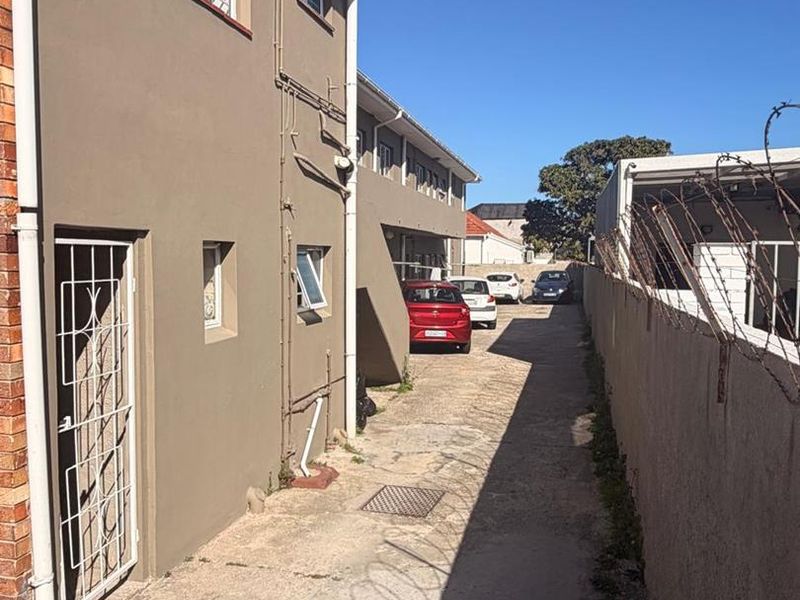 A side exterior view of the apartment building showing a concrete driveway, security fencing, and access to the rear area.