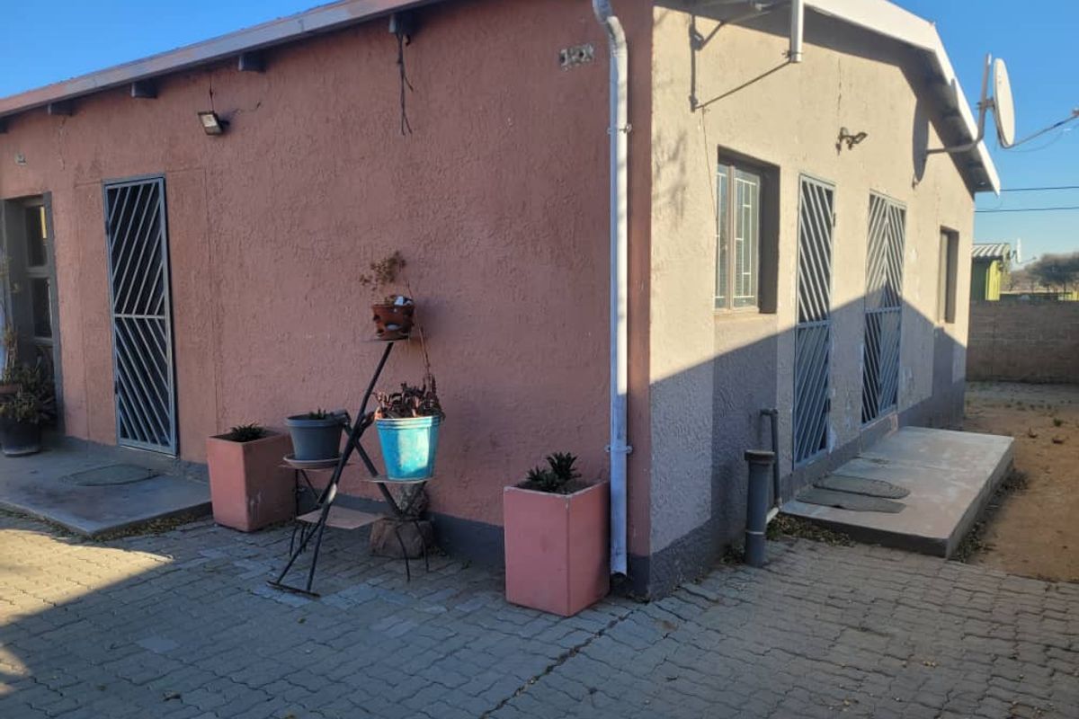 Rear exterior view of the property featuring paved ground, security gates on doorways, and external wall finishes showing signs of wear and weathering.