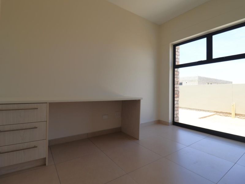 A bright, modern home office featuring neutral tiled flooring, a built-in desk, and a large window providing ample natural light.