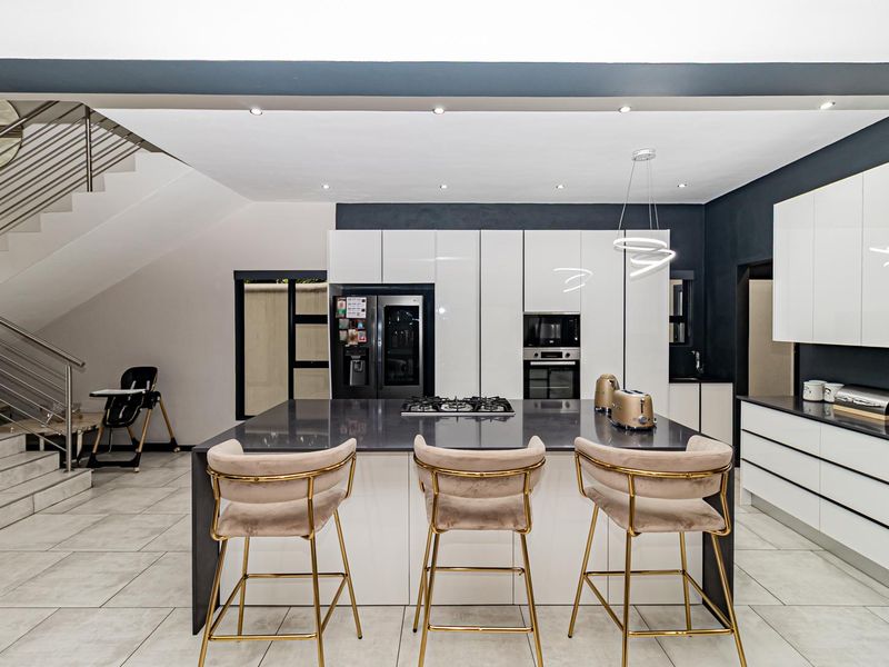 Modern kitchen featuring a central island with gas hob, high-gloss cabinetry, stone countertops, and an adjacent staircase with a sleek stainless steel balustrade.