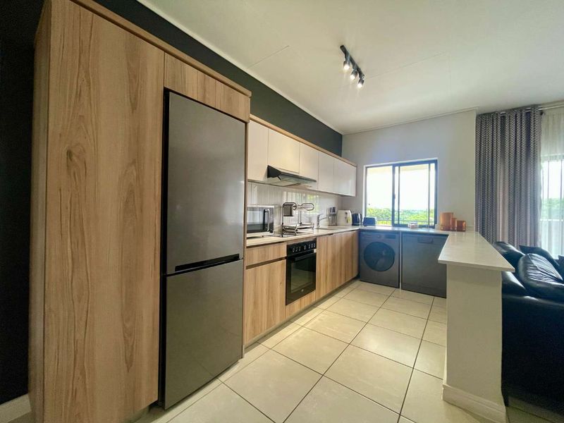 Modern open-plan kitchen featuring wood-finish cabinetry, integrated appliances, tiled flooring, and a breakfast bar overlooking the living area.