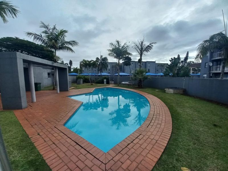 Communal outdoor pool area featuring a swimming pool, brick paved surround, covered patio structure, and landscaped garden within an apartment complex.