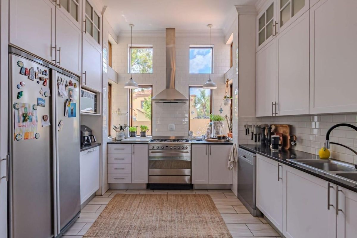 A bright, modern galley-style kitchen featuring white cabinetry, subway tile backsplash, stainless steel appliances, and large windows providing natural light.