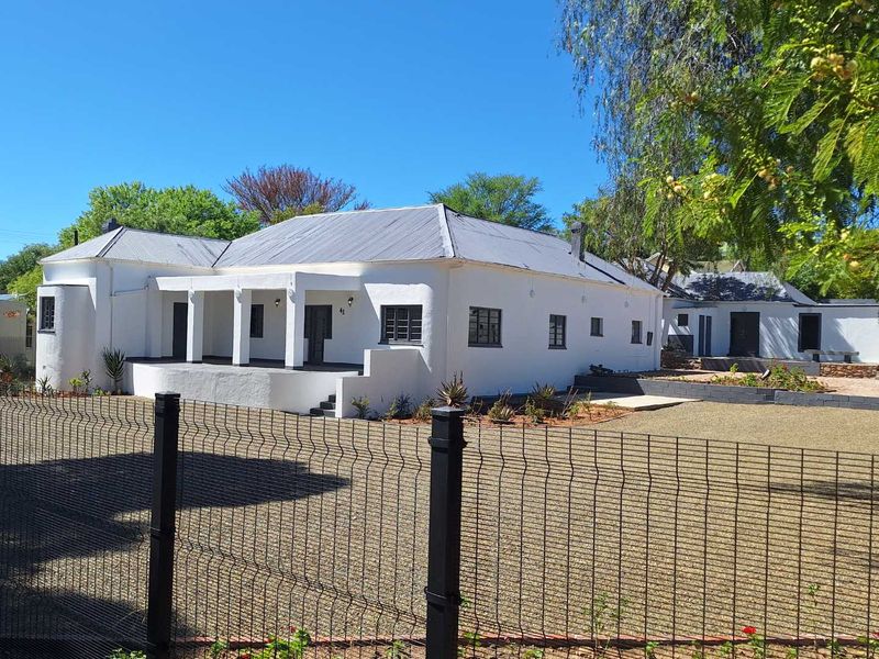 A well-maintained guest house exterior featuring a white facade, corrugated metal roof, and a spacious covered porch, set behind a secure mesh fence.
