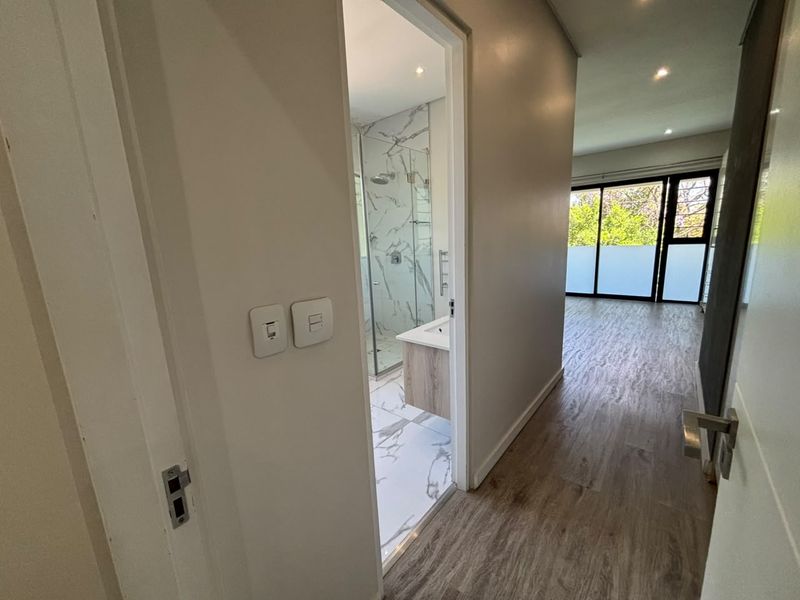 A bright hallway leading to a modern bathroom with marble-look tiling and a glass shower, adjacent to an open-plan living area with sliding doors.