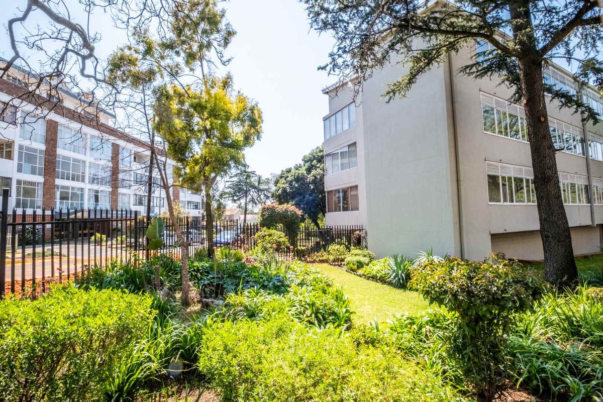 A well-maintained communal garden area situated between multi-story apartment buildings, featuring mature trees, lush greenery, and secure metal perimeter fencing.