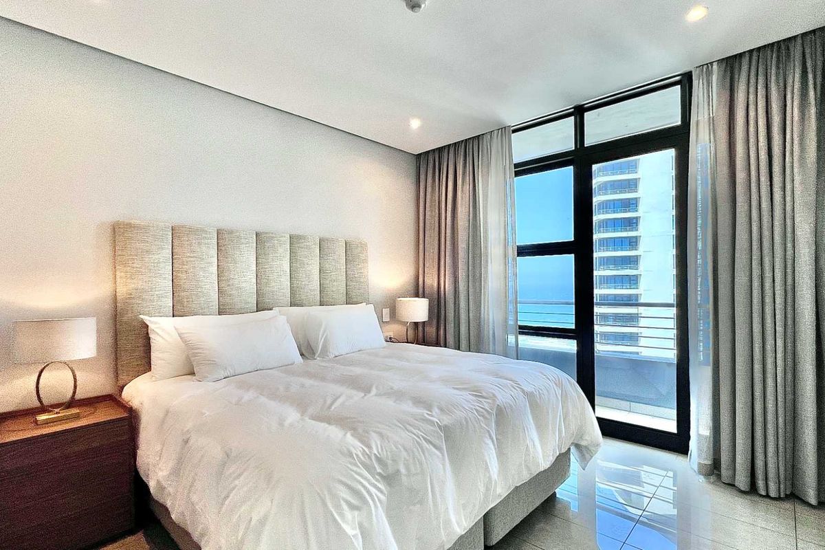 Modern bedroom featuring floor-to-ceiling windows with balcony access, neutral finishes, and recessed lighting, offering a bright and contemporary living space.