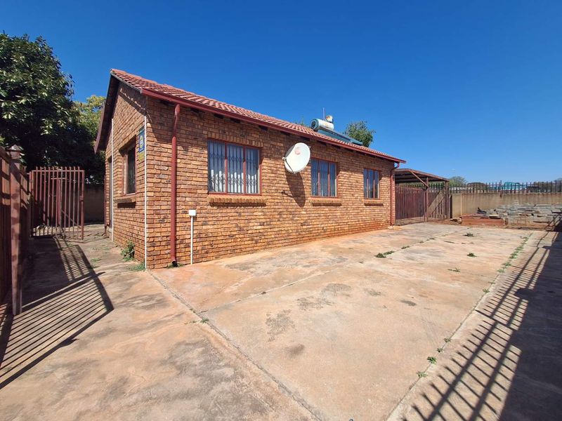 Rear view of a face brick house featuring a tiled roof, solar geyser, security bars, and a large concrete driveway with a carport.