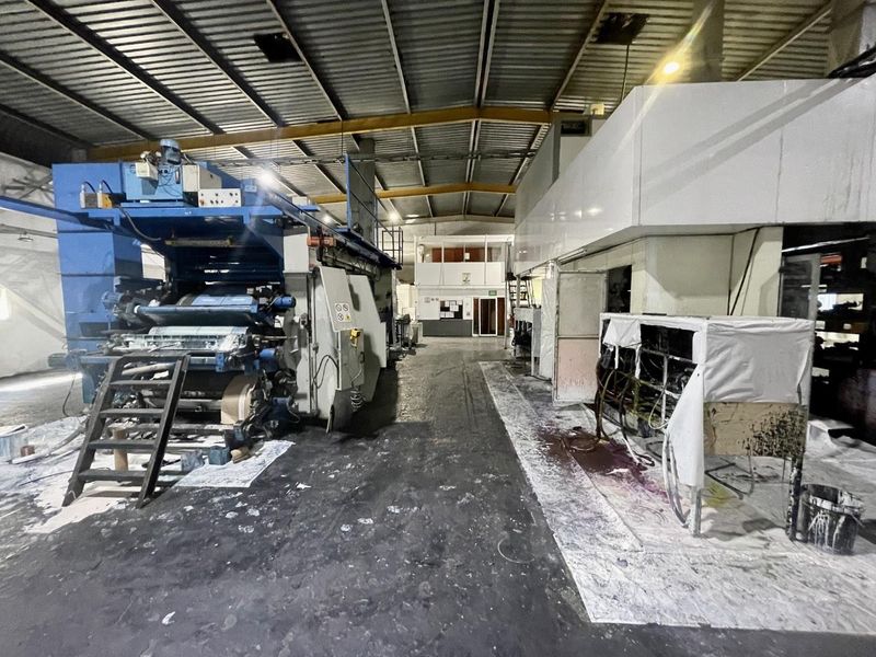 Industrial warehouse space featuring heavy-duty printing machinery, exposed steel roof trusses, and a mezzanine office level.