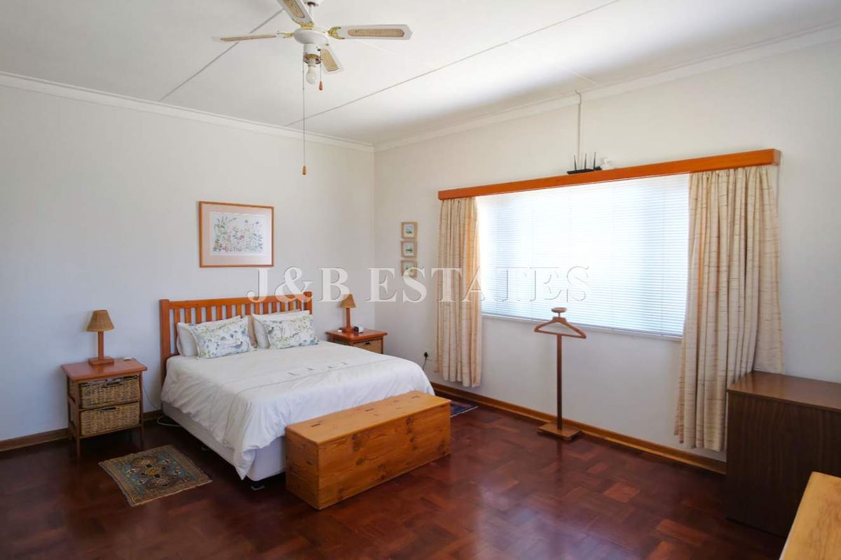 A spacious bedroom featuring classic parquet flooring, a ceiling fan, and large windows with fitted blinds, offering a neutral and functional living space.