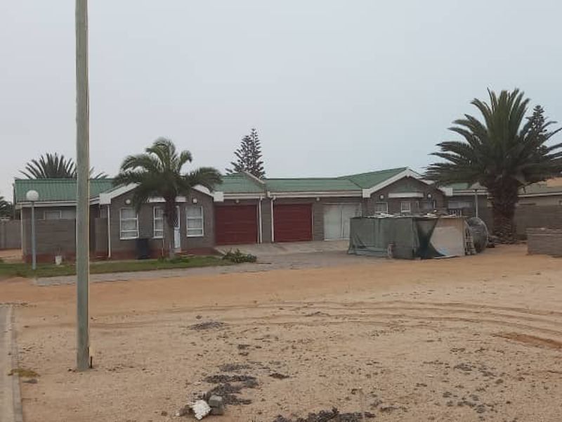 Front view of a single-story house featuring a double garage, metal roofing, and a perimeter wall, set on a large sandy plot.