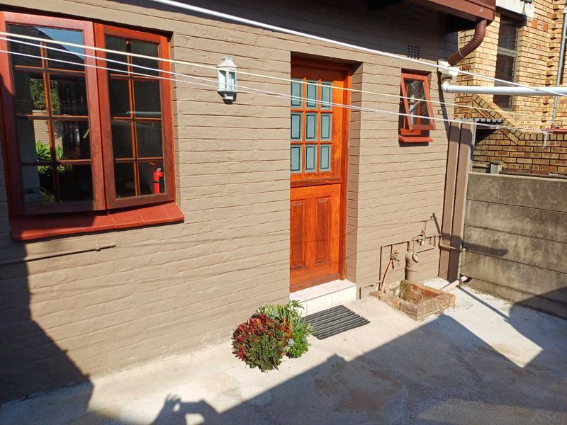 Rear exterior view of the cottage featuring timber-framed windows, a wooden entrance door, and a concrete patio area with external plumbing fixtures.
