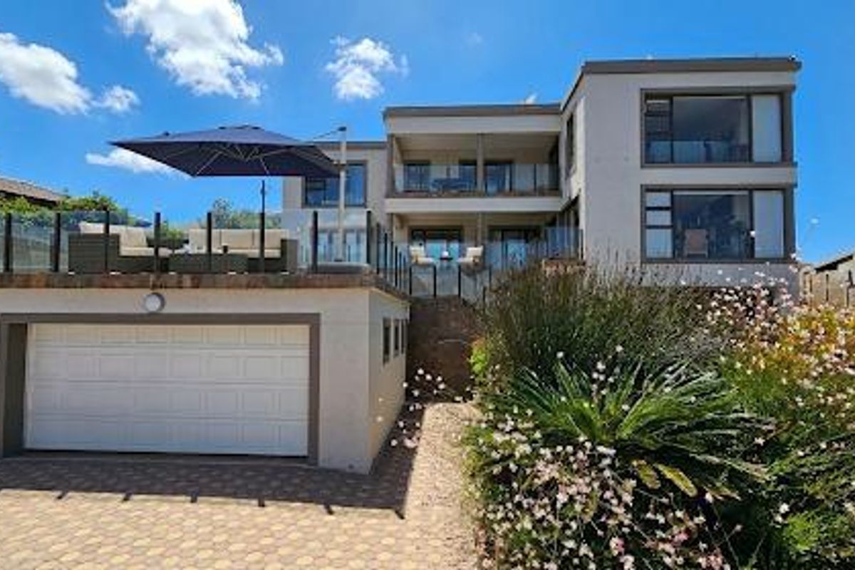Rear view of a multi-level modern home featuring a raised patio with glass balustrades, a double garage, and landscaped garden area.