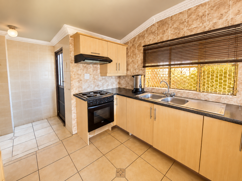 A functional kitchen featuring light wood cabinetry, a built-in oven with hob, tiled splashbacks, and a double sink with a view through security-screened windows.