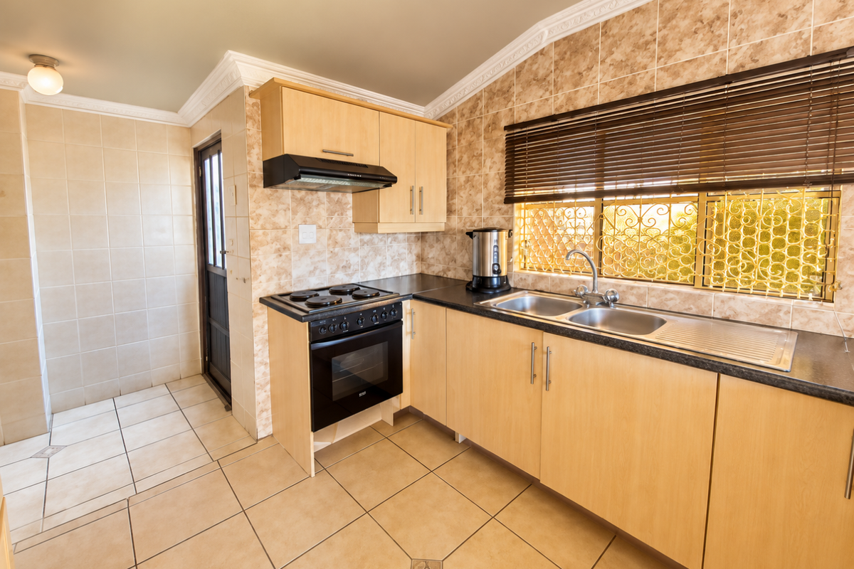 A functional kitchen featuring light wood cabinetry, a built-in oven with hob, tiled splashbacks, and a double sink with a view through security-screened windows.