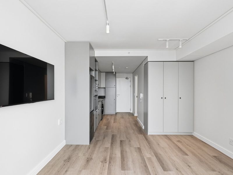 Modern open-plan living area featuring light wood-look laminate flooring, sleek grey built-in cabinetry, integrated kitchen appliances, and minimalist track lighting.