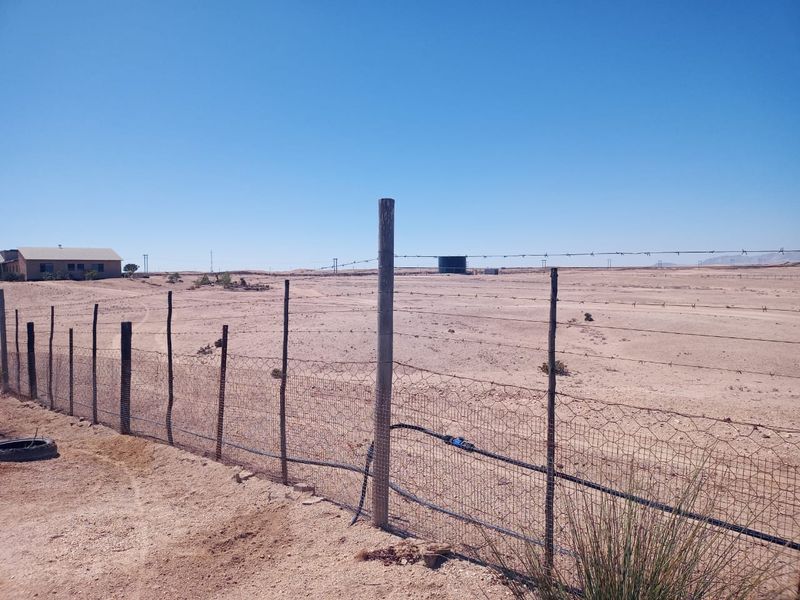 Expansive arid small holding land featuring perimeter wire fencing, irrigation infrastructure, and a distant water storage tank under a clear sky.