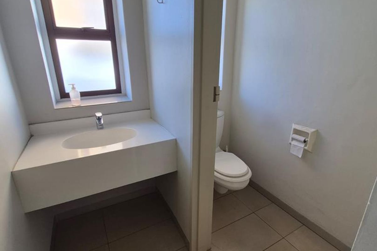 A clean and functional office bathroom featuring a vanity basin, toilet, and neutral floor tiling with natural light from a frosted window.