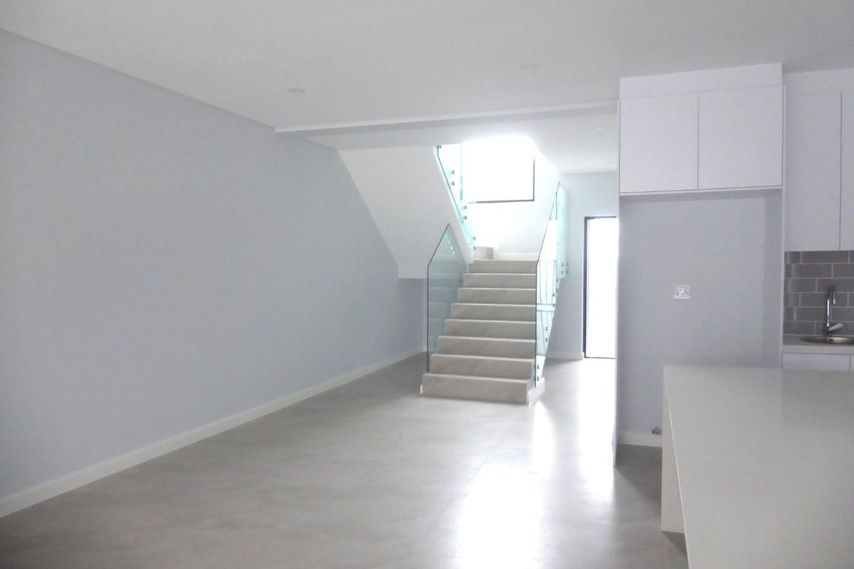 Modern open-plan living area featuring a sleek staircase with glass balustrades, neutral tiled flooring, and a contemporary kitchen with white cabinetry.