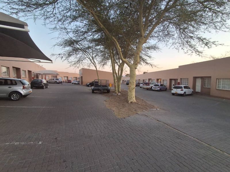 Exterior view of the townhouse complex featuring a paved driveway, designated parking bays, and mature trees providing shade to the residential units.