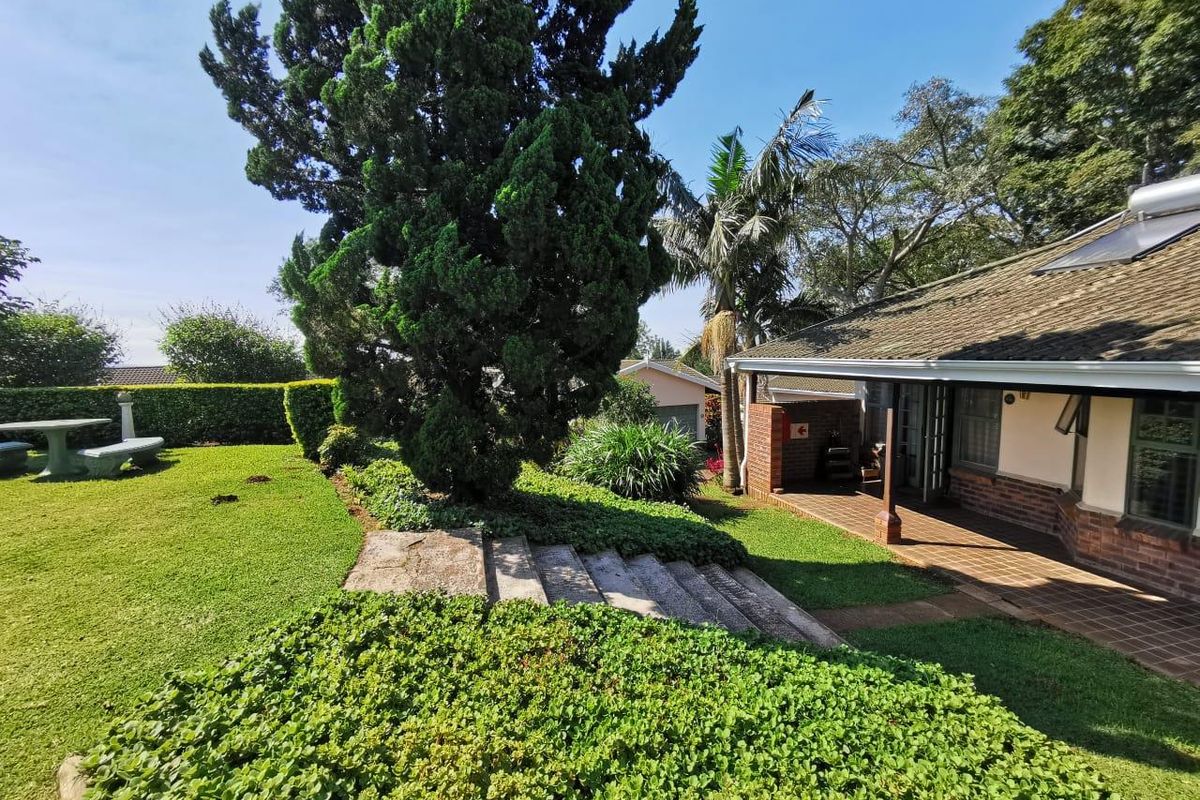 A well-maintained rear garden featuring a covered tiled patio, brick exterior walls, and stone steps leading to a lush lawn area.