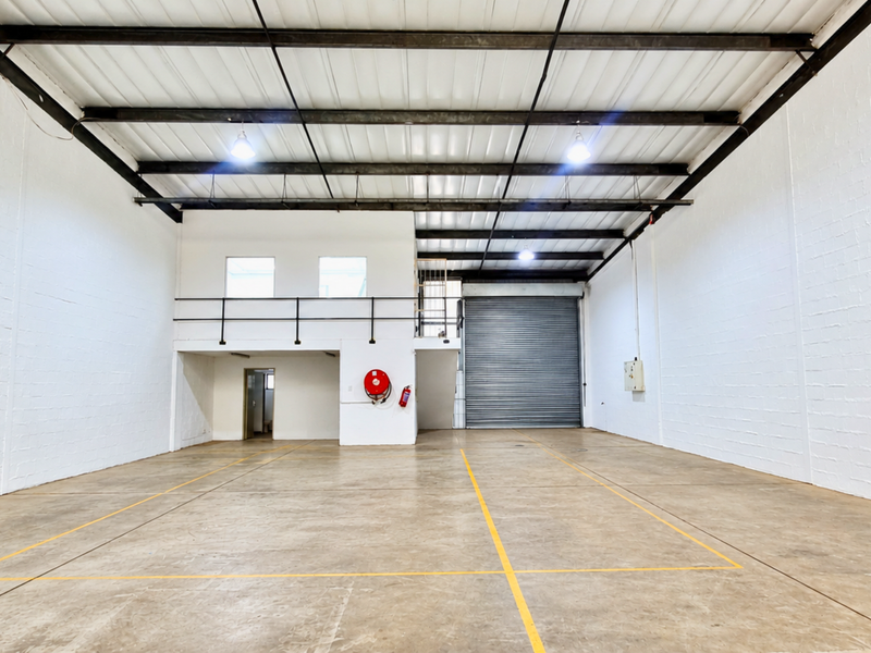Spacious industrial warehouse unit featuring high ceilings with exposed steel trusses, a large roller shutter door, and a functional mezzanine level.
