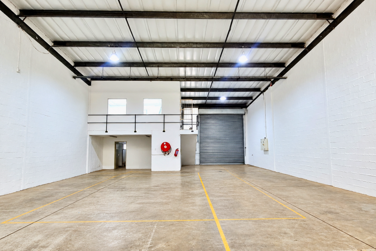 Spacious industrial warehouse unit featuring high ceilings with exposed steel trusses, a large roller shutter door, and a functional mezzanine level.