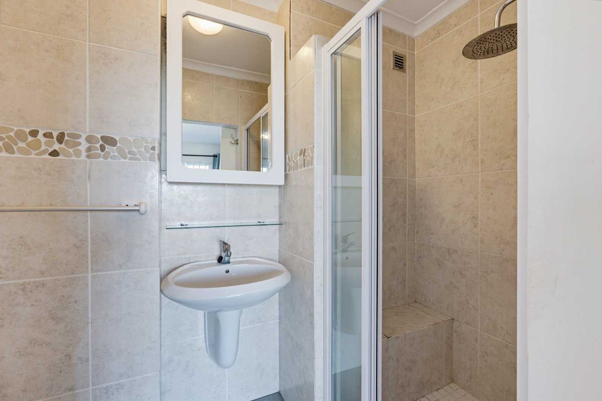 A clean, functional bathroom featuring a pedestal washbasin, wall-mounted mirror, and a glass-enclosed shower with a built-in bench and rainfall shower head.