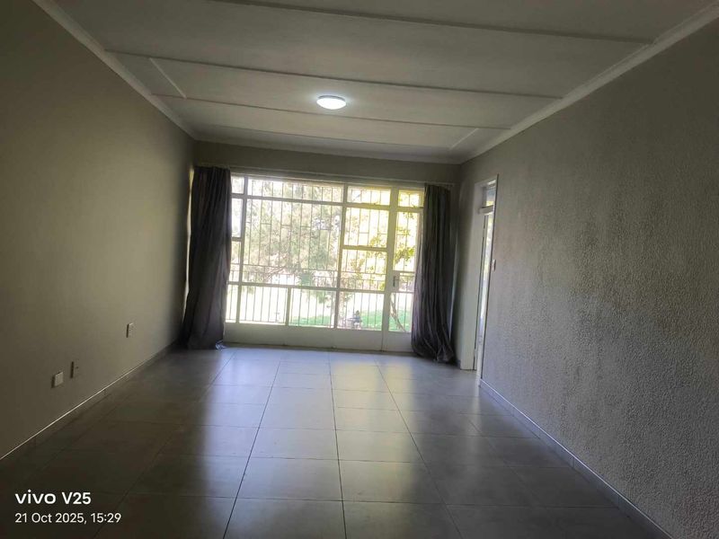 A spacious, empty living room featuring neutral tiled flooring, textured walls, and a large window allowing for natural light.