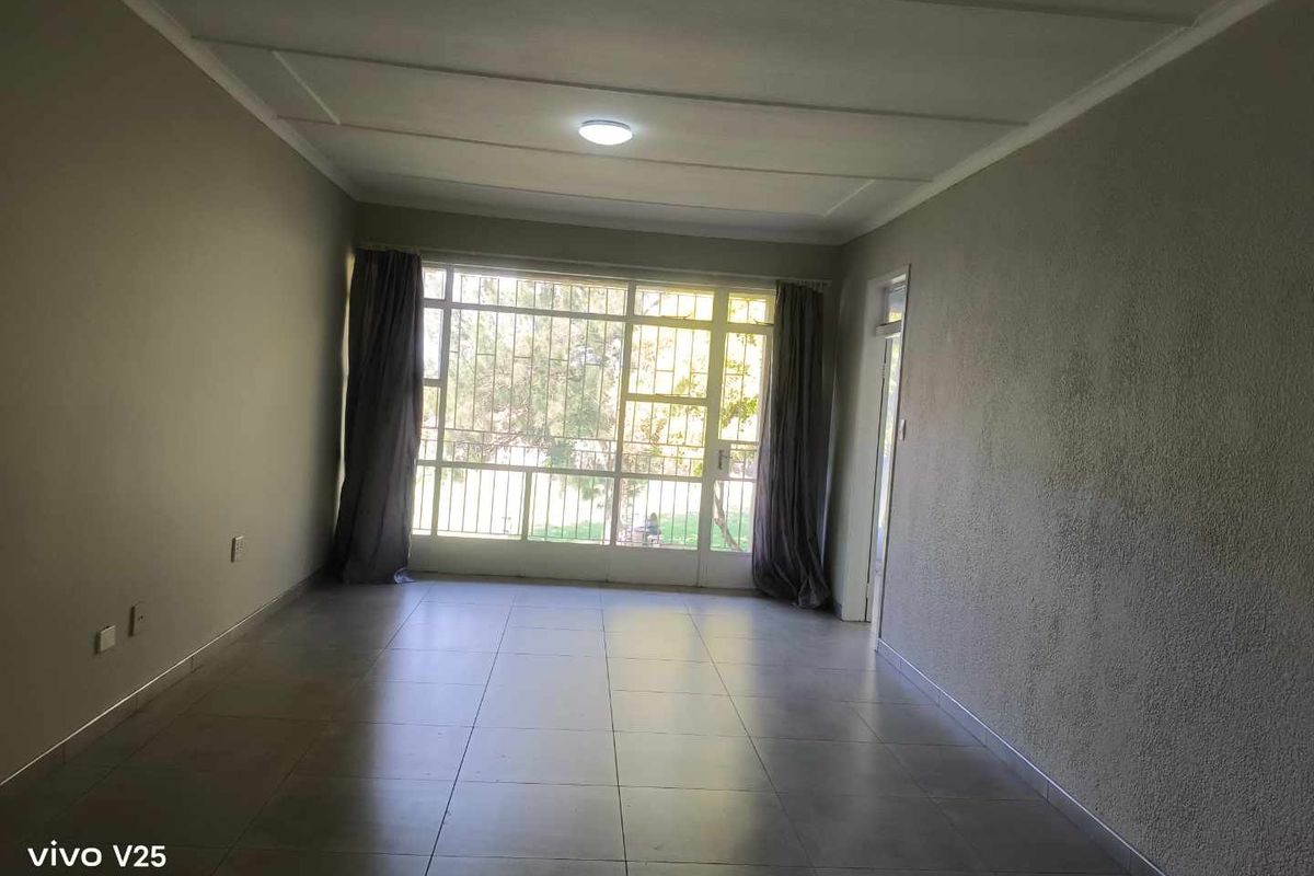 A spacious, empty living room featuring neutral tiled flooring, textured walls, and a large window allowing for natural light.