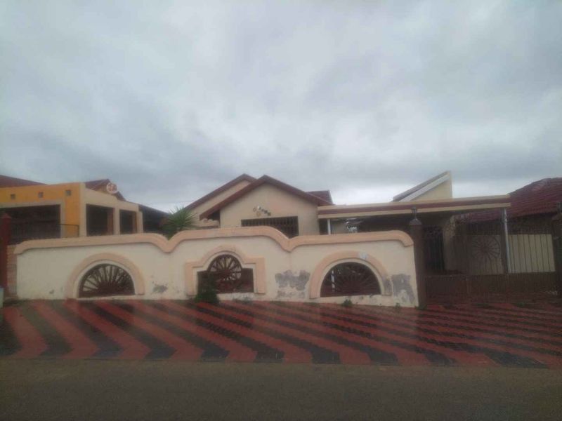 Front elevation of a single-story house featuring a decorative boundary wall, paved driveway, and gated entrance under a cloudy sky.
