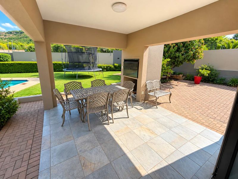 A spacious covered patio featuring tiled flooring and a built-in braai, overlooking the garden and pool area.