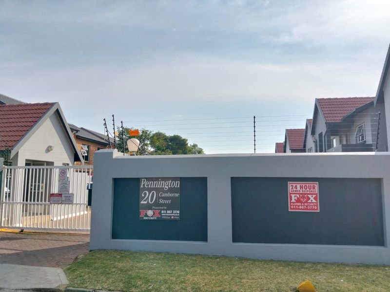 Front entrance view of the townhouse complex featuring a secure gated access, boundary wall with electric fencing, and paved driveway.