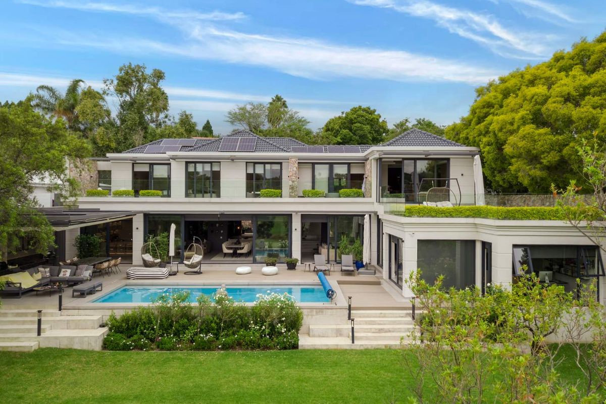 A spacious rear exterior view of a modern two-story home featuring a swimming pool, expansive decking, glass balustrades, and solar panels on the roof.