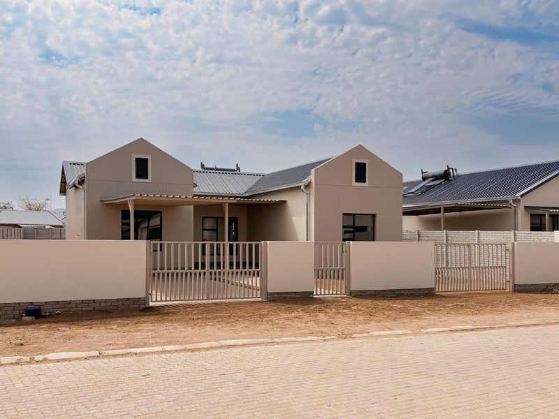 Modern single-story house featuring a metal roof, solar water heater, secure perimeter wall with metal gates, and a paved driveway.